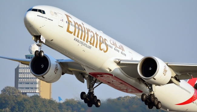 An Emirates Boeing 777-300ER aircraft on takeoff with landing gear extended and a control tower in the background