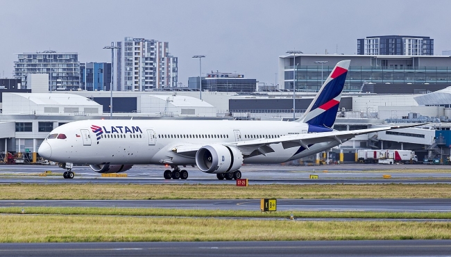 A LATAM Boeing 787 Dreamliner jet aircraft landing on a runway at an airport with buildings in the background